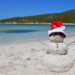 A sand-snowman wearing a red Santa hat and sunglasses, sitting on a sunlit white beach with clear blue water and a green forested shoreline in the background.