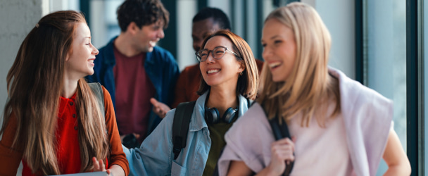 Happy university students walk and laugh together in a bright library hallway, carrying backpacks, laptops and books while sharing friendship, collaboration and youthful energy.