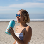 Woman wearing sunglasses drinking from a blue water bottle at the beach on a hot summer day, representing staying hydrated during Australian summer.