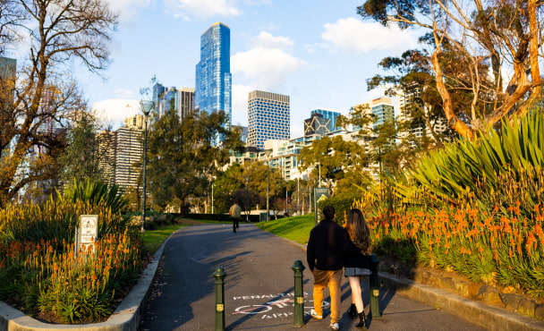 Couple walking into Flagstaff Gardens in Melbourne on treelined path and city skyline in the background.