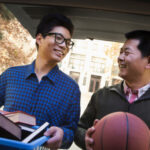 Father and son in back of car in front of dormitory. Father is helping son move in. They are carrying books and a basketball.