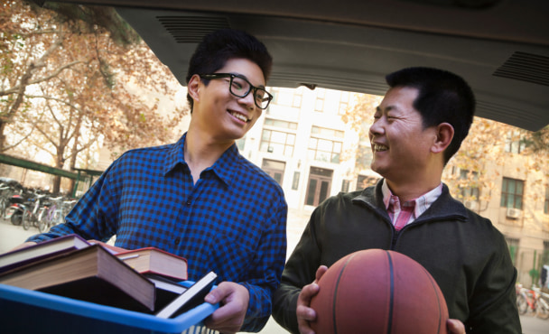 Father and son in back of car in front of dormitory. Father is helping son move in. They are carrying books and a basketball.