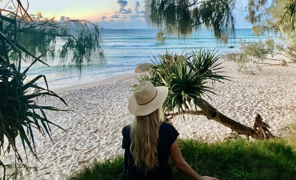 Horizontal seascape of woman in woven hat sitting in shady green grass between tropical pandanus trees looking out to ocean views.