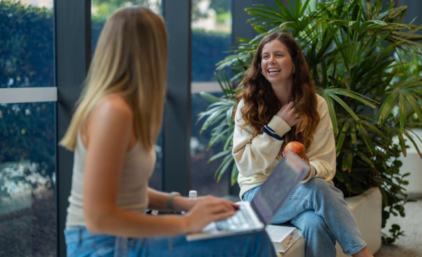 Two young women sit together in a university campus library in Australia, studying and discussing content from a textbook and laptop.