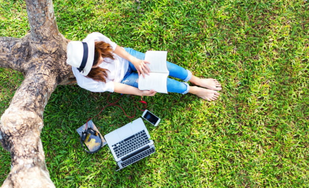 Girl enjoy listening music and reading a book and play laptop on the grass field of the park in the morning.