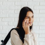 Uni student talking on mobile phone to parent, carrying backpack on shoulder with a white brick wall background.