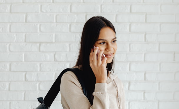Uni student talking on mobile phone to parent, carrying backpack on shoulder with a white brick wall background.