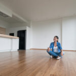 A woman sitting on the floor of a bright, modern unfurnished apartment with wooden floors and large windows, illustrating the concept of furnished vs unfurnished student accommodation.