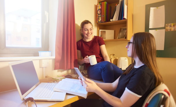Two female university students sitting at a desk in a dorm room relaxing and having a chat and a cup of tea.