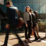 Businesswoman holding her hand bag standing still and looking to the sky on a busy street with blurred business people walking past her.