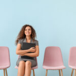 Young woman holding a black folder, waiting for job interview, sitting on a row of pink seats against a blue background.