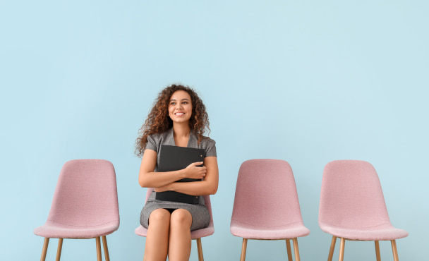 Young woman holding a black folder, waiting for job interview, sitting on a row of pink seats against a blue background.