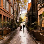 A couple of people walking down a street next to tall buildings in Melbourne laneway with autumn leaves on the ground.