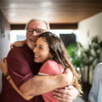 Granddaughter embracing grandfather at student accommodation, with grandmother in background.