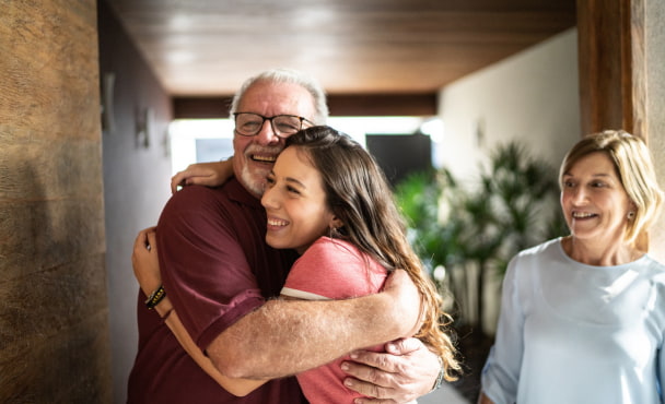 Granddaughter embracing grandfather at student accommodation, with grandmother in background.