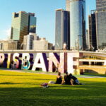 People sitting on green grass field in front of Brisbane sign, near high rise buildings during daytime.