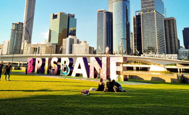 People sitting on green grass field in front of Brisbane sign, near high rise buildings during daytime.