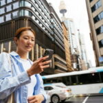 Young Asian woman using public transport in the city with cars and a bus behind her. The Sydney Tower Eye and office buildings are in the background.