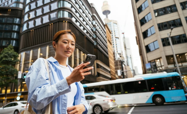 Young Asian woman using public transport in the city with cars and a bus behind her. The Sydney Tower Eye and office buildings are in the background.