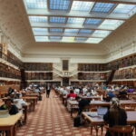 Inside a large, ornate library with soaring ceilings with light coming through, filled with large desks and people studying, and lots of books on the walls.
