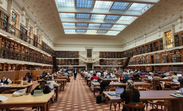 Inside a large, ornate library with soaring ceilings with light coming through, filled with large desks and people studying, and lots of books on the walls.
