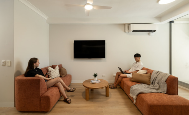 Modern student accommodation living room with two students on red lounges with a coffee table in between and a television on the wall.