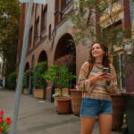 Young woman smiling while walking on a city sidewalk holding a mobile phone and exploring the student accommodation neighbourhood in Australia