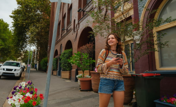 Young woman smiling while walking on a city sidewalk holding a mobile phone and exploring the student accommodation neighbourhood in Australia