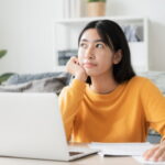 Asian student sitting in front of laptop at home contemplating when to book student accommodation online.