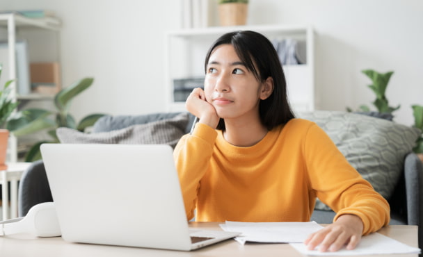 Asian student sitting in front of laptop at home contemplating when to book student accommodation online.