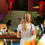 Woman in floral dress ordering at a snack bar with Asian decor and light refreshments visible.