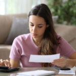 Woman doing accounting holding bill and using calculator on a coffee table at home.