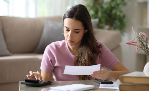 Woman doing accounting holding bill and using calculator on a coffee table at home.